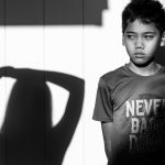 Portrait of a child standing in front of a wall and looking towards the source of a shadow on the wall next to the child.