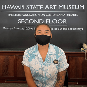 Person seated in front of a large sign that reads "Hawaii State Art Museum".