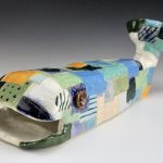 Ceramic whale with surface texture and glaze colors that look like a patchwork of different pieces sewn together. The visible eye is shaped like a brown button. The mouth of the whale is open and the tail is lifted up.