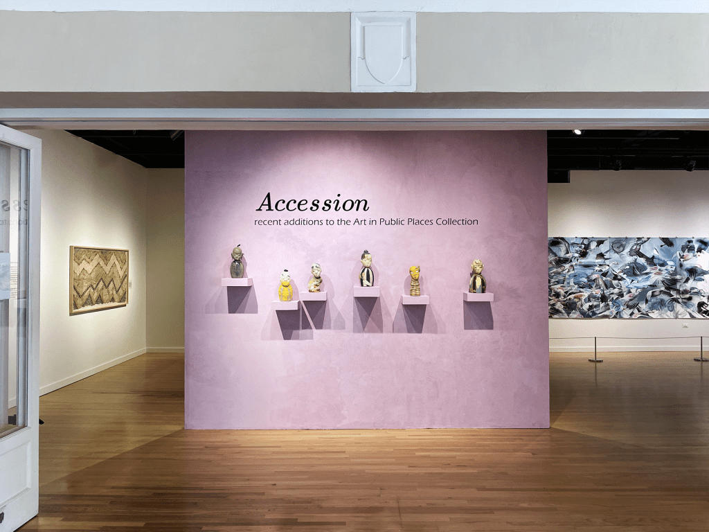View of museum gallery entrance, with a light purple wall holding six small sculptures. A framed kapa in shades of brown and green is visible on the left, on a separate wall, and part of a large blue, white, and black drawing is visible on the right side on a back wall.