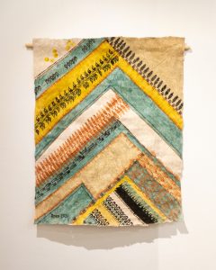 Rectangle of kapa painted and printed with triangles and chevron shapes evoking mountain peaks. Colors are shades of yellow, green, brown, and black.