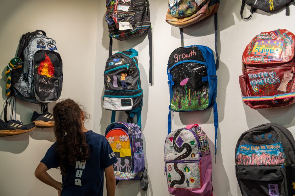 Child looks up at a wall where several art backpacks are on display.