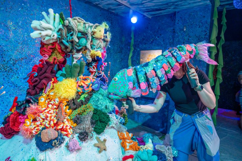 Puppeteer holding large parrotfish puppet up to colorful display of coral made of yarn