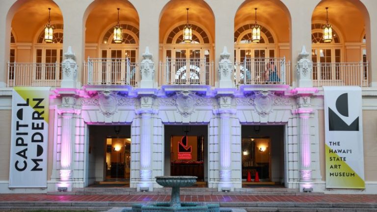 View of entrance to No. 1 Capitol District Building, lit with pink and purple lights. Banners on either side of the entrance read 'Capitol Modern' and 'The Hawaii State Art Museum'.