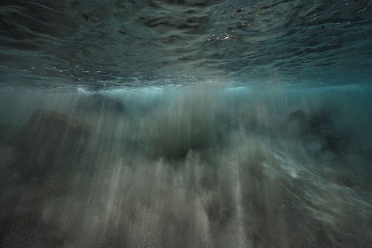 underwater photograph of light streaming down and clouds of sand rising up.