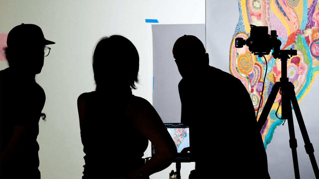 Three people examining a photo preview of an artwork being photographed.