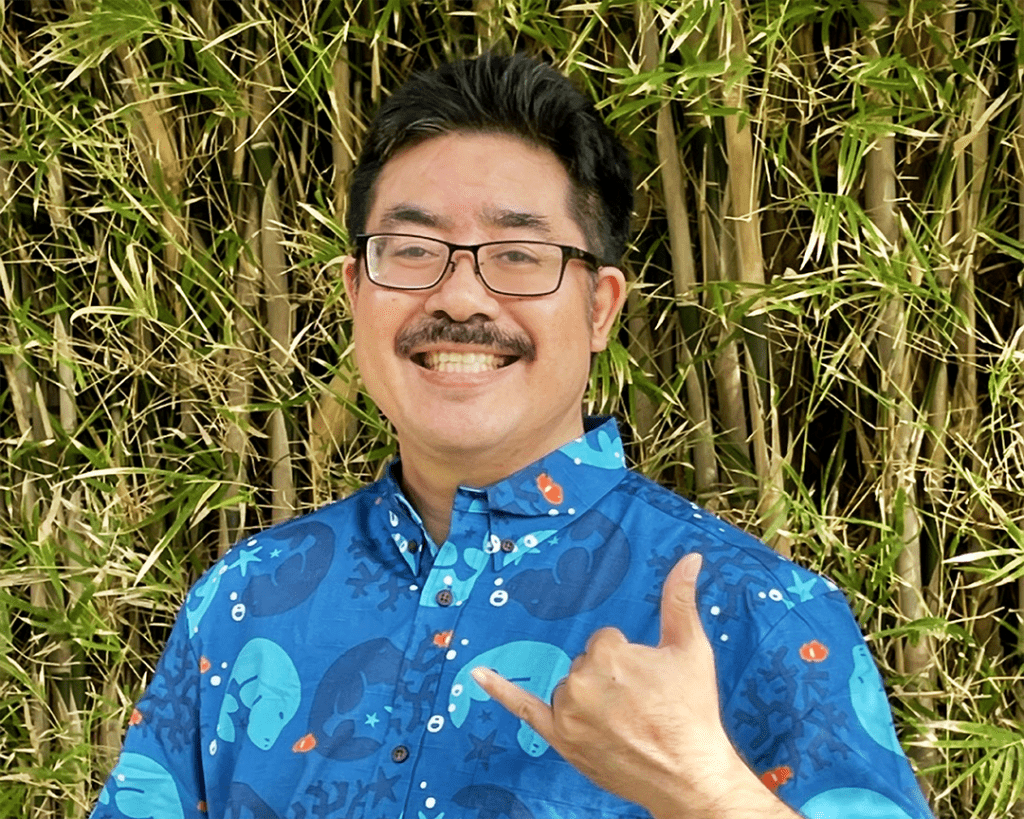 Lee Tonouchi smiling at the camera and throwing a shaka sign with his left hand. Lee is wearing glasses and a bright blue aloha print shirt.